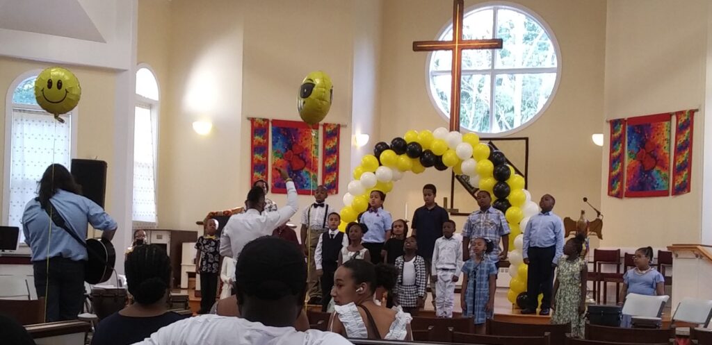 Children from Rose Conservatory sing at the front of a church with a yellow and black balloon arch. A guitarist stands to the left playing, while a director conducts. The bright church sanctuary features colorful banners and large windows.