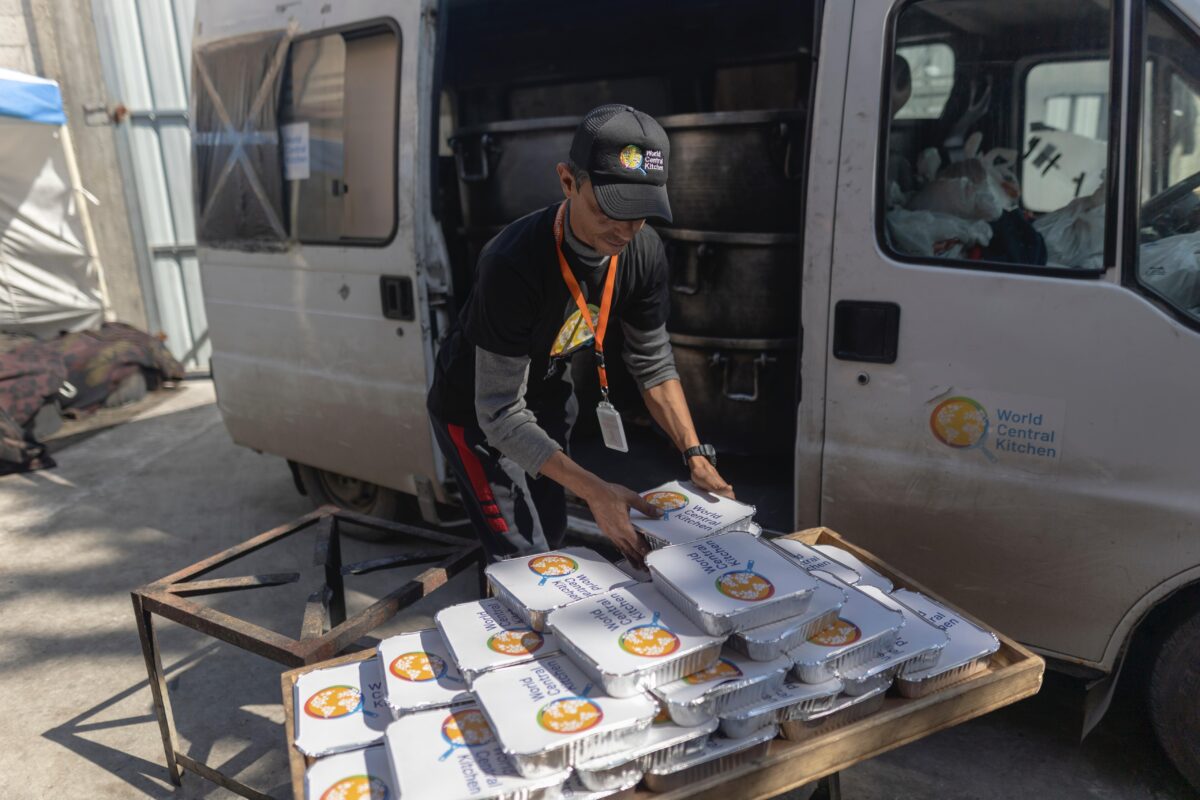 An aid worker unloads meals packaged in foil trays from a van. The meals have covers with the World Central Kitchen logo.