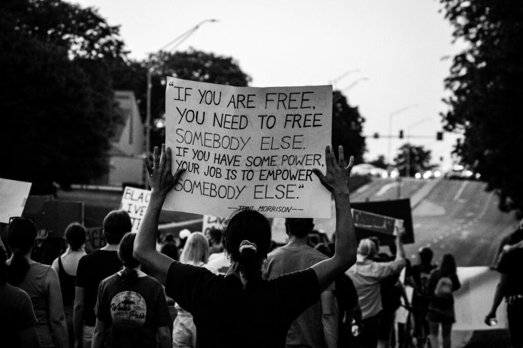 This black-and-white photograph captures a protest scene with a group of people marching down a street. The focal point is a person in the foreground holding up a large sign that reads:

"IF YOU ARE FREE, YOU NEED TO FREE SOMEBODY ELSE. IF YOU HAVE SOME POWER, YOUR JOB IS TO EMPOWER SOMEBODY ELSE."
—Toni Morrison